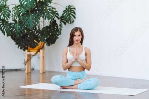 Attractive young woman wearing sport bra sit in lotus yoga position in light room. Relaxing state of mind, meditation, namaste