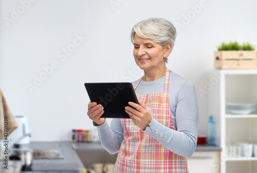 cooking, culinary and old people concept - happy smiling senior woman in apron with tablet pc computer over home kitchen background