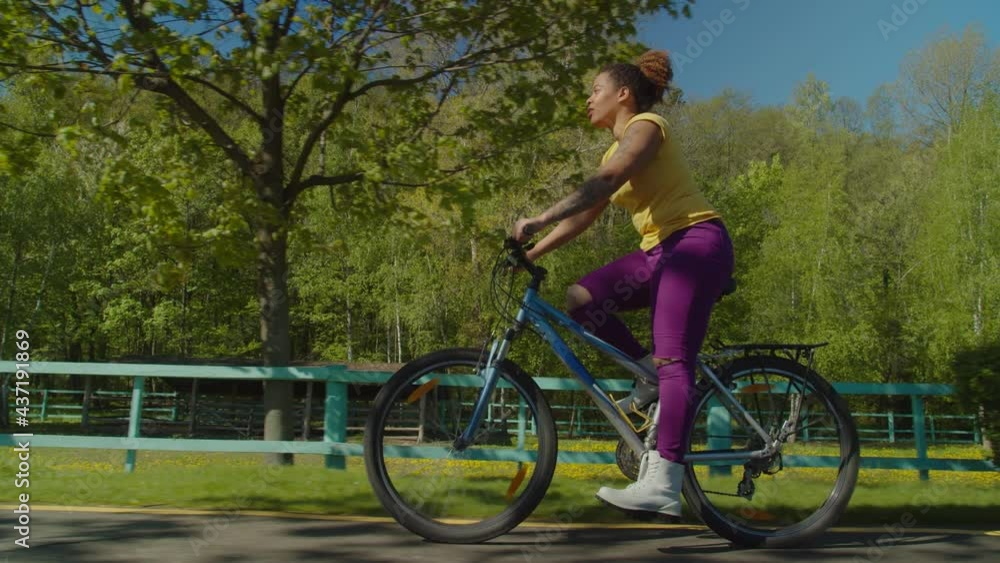 Side view of attractive positive hipster african woman riding on bicycle in public park, enjoying active healthy lifestyle, freedom and beautiful summer nature while cycling outdoors in early morning Side view of attractive positive hipster african woman riding on bicycle in public park, enjoying active healthy lifestyle, freedom and beautiful summer nature while cycling outdoors in early morning