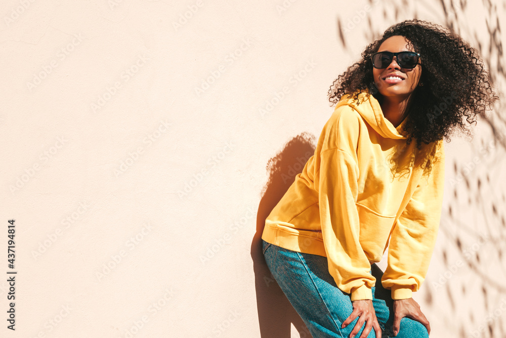 Fototapeta premium Beautiful black woman with afro curls hairstyle.Smiling hipster model in yellow hoodie. Sexy carefree female posing on the street background in sunglasses. Cheerful and happy