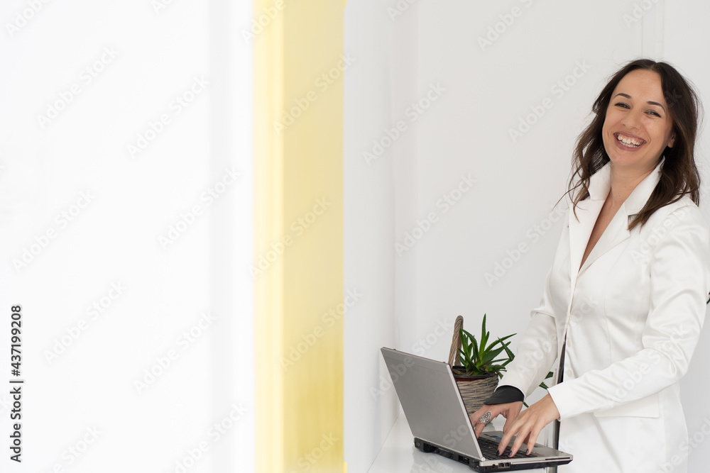 Elegantly dressed smiling young woman in a white blazer and black pants stands next to a desk on which is a laptop and working focused on the computer. White background and flowers plant on the table Elegantly dressed smiling young woman in a white blazer and black pants stands next to a desk on which is a laptop and working focused on the computer. White background and flowers plant on the table
