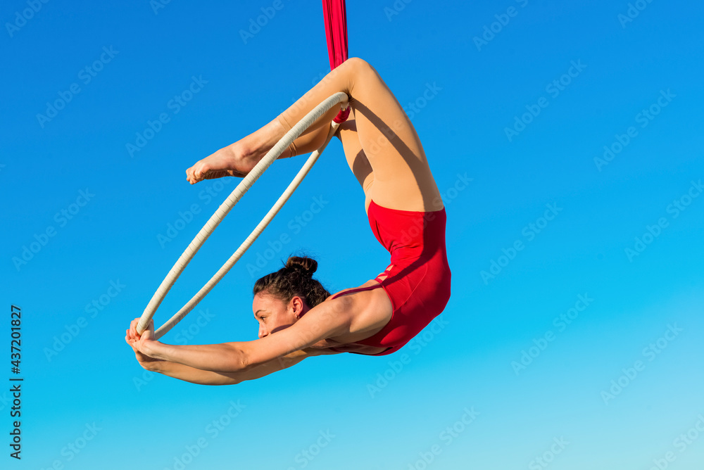 acrobat athletic, young graceful gymnast performing aerial exercise in the air ring outdoors on sky background. flexible woman in red suit performs circus artist dancing on hoop demonstrates poses. - obrazy, fototapety, plakaty 