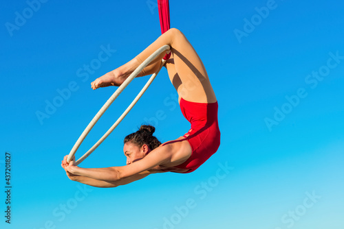 acrobat athletic, young graceful gymnast performing aerial exercise in the air ring outdoors on sky background. flexible woman in red suit performs circus artist dancing on hoop demonstrates poses. photo