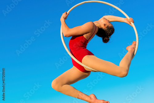 acrobat athletic, young graceful gymnast performing aerial exercise in the air ring outdoors on sky background. flexible woman in red suit performs circus artist dancing on hoop demonstrates poses.