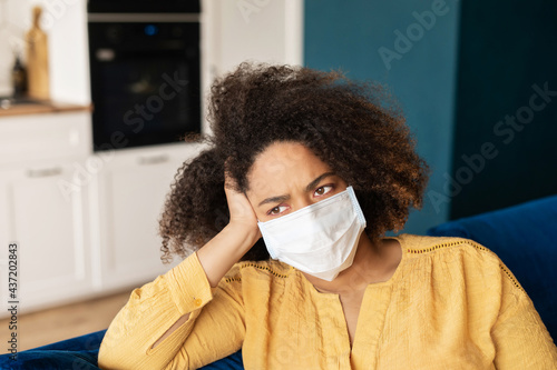 Frustrated young woman wearing medical mask sits on the couch at home alone, upset African woman feels lonely during self isolation, female worrying about covid pandemic