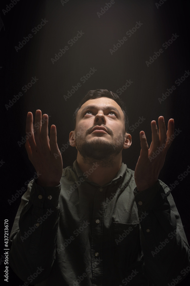 Portrait of a young man raising his hands in a supplicating prayer gesture and looking up at a descending stream of light on a dark background.