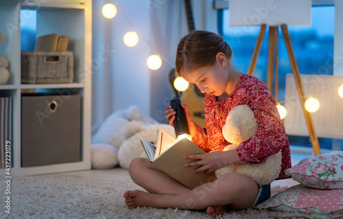 Little beautiful girl reads a fairy tale before slleping with flashlight and Teddy bear. Child sitting in the children bedroom with book. Evening reading with garland photo