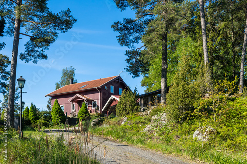 Traditional Swedish wooden house near forest. Countryside village in Scandinavia. Red typical cottage rural house with porch. Carport. In courtyard Landscaping of garden with ornamental thuja plants.