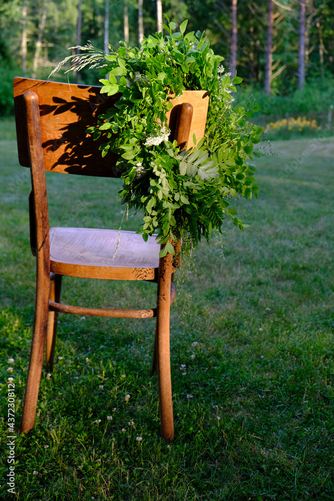 Green leaf and flower solstice wreath sunlit, hanging on the back of an antique wooden chair, placed in a green meadow