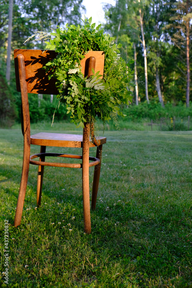 Green leaf and flower solstice wreath sunlit, hanging on the back of an antique wooden chair, placed in a green meadow