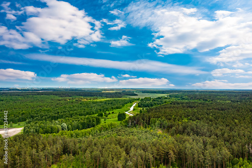 Aerial panoramic view of nature. The road in the fields and dense forest. Fields  meadows  road  outside urban nature in Europe. Blue beautiful sky and white clouds. Natural landscape  summer. 