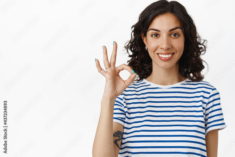 Portrait of brunette girl with white perfect smile, shows okay OK gesture, approve and like, agree or say yes, standing in t-shirt against white background Portrait of brunette girl with white perfect smile, shows okay OK gesture, approve and like, agree or say yes, standing in t-shirt against white background