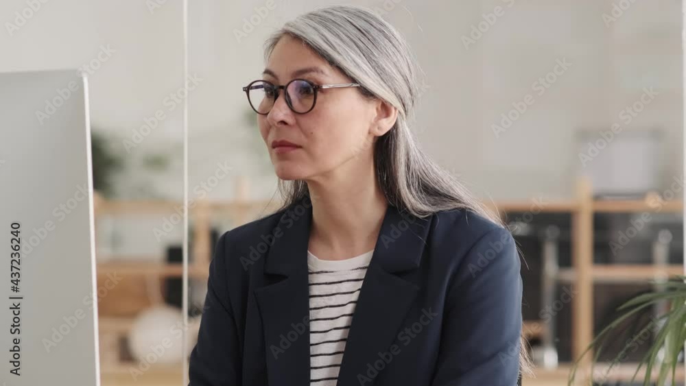 A confident asian woman with gray hair looking in surprise at the monitor and taking off her glasses while sitting at a table in the office A confident asian woman with gray hair looking in surprise at the monitor and taking off her glasses while sitting at a table in the office