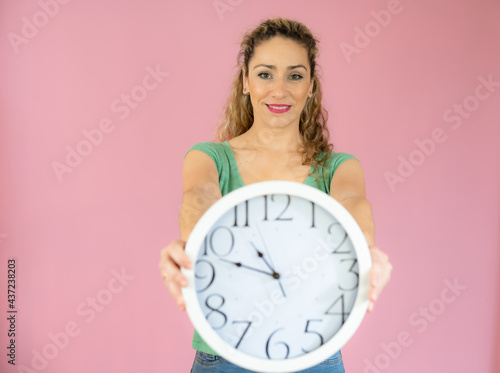 Portrait of a shocked woman in casual t-shirt holding alarm clock isolated over pink background