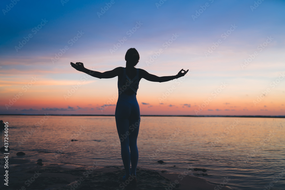 Back silhouette view of calm female yogi with raised hands getting energy from nature during evening meditation at coastline seashore, tranquil woman in tracksuit enjoying own harmony during sunset