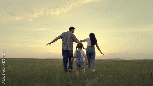 A happy family runs across the field with a small child at sunset in the sky, running mom, dad and kid are jumping cheerfully in the field in the evening, the team is traveling cheerfully playing
