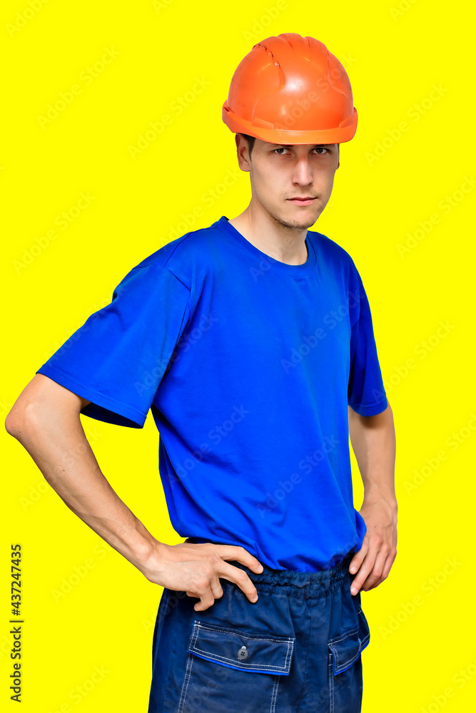 Studio portrait of a confident worker in a construction helmet looking at the camera and holding his hands on his belt. A young guy of Caucasian appearance is isolated on a yellow background. Studio portrait of a confident worker in a construction helmet looking at the camera and holding his hands on his belt. A young guy of Caucasian appearance is isolated on a yellow background.