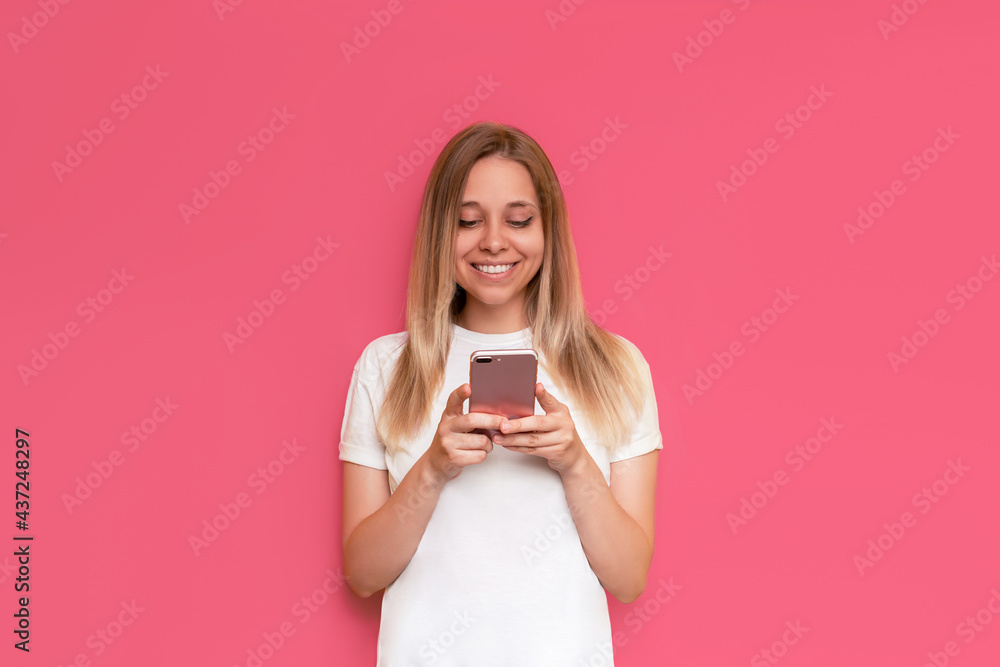 A young caucasian happy pretty blonde woman smiles and holds a mobile phone looking at the screen isolated on bright color pink background. The girl in a white t-shirt uses her smartphone. Copy space A young caucasian happy pretty blonde woman smiles and holds a mobile phone looking at the screen isolated on bright color pink background. The girl in a white t-shirt uses her smartphone. Copy space