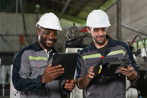 Two male engineers happy to work holding tablet and remote control for control a robot arm welding machine in an industrial factory.