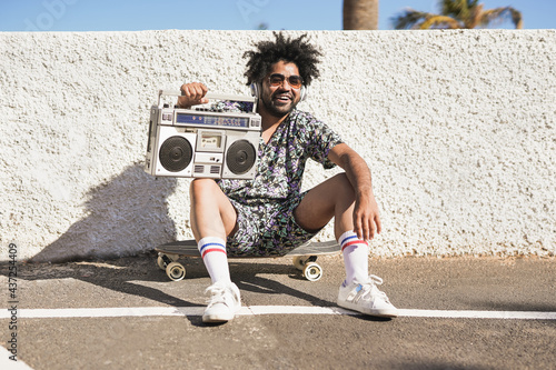 Young african man listening music playlist while sitting on skateboard outdoor - Skater boy with vintage boombox in the city photo