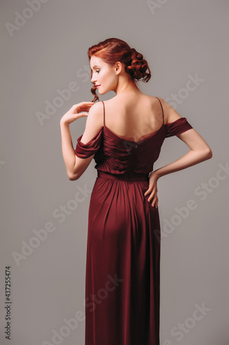 Beautiful skinny ginger girl in a burgundy evening gown. Maroon red dress with off the shoulder sleeves. Studio portrait of young lady on grey background.