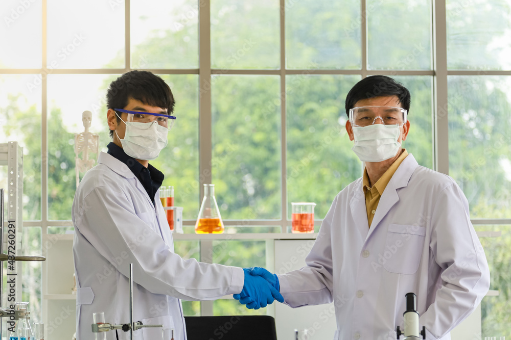 Portrait of professional senior and young doctor scientist wearing lap safety glasses, surgical face mask and blue rubber glove make a hand shake and looking to camera in working laboratory. Portrait of professional senior and young doctor scientist wearing lap safety glasses, surgical face mask and blue rubber glove make a hand shake and looking to camera in working laboratory.