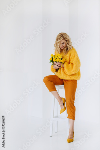 Happy young blonde woman with a bouquet of yellow roses in a voluminous sweater posing on a white background in the studio. Soft selective focus.