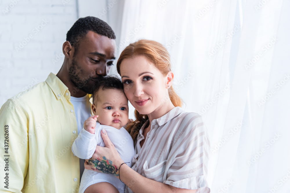 happy woman looking at camera near smiling husband kissing head of infant daughter happy woman looking at camera near smiling husband kissing head of infant daughter