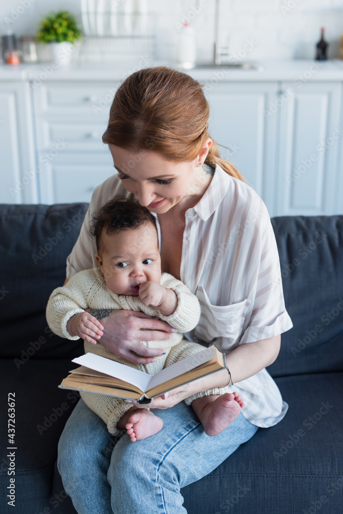 woman reading book while sitting on sofa with infant african american kid woman reading book while sitting on sofa with infant african american kid