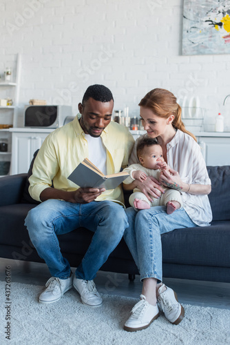 smiling woman sitting on sofa with baby near african american husband reading book