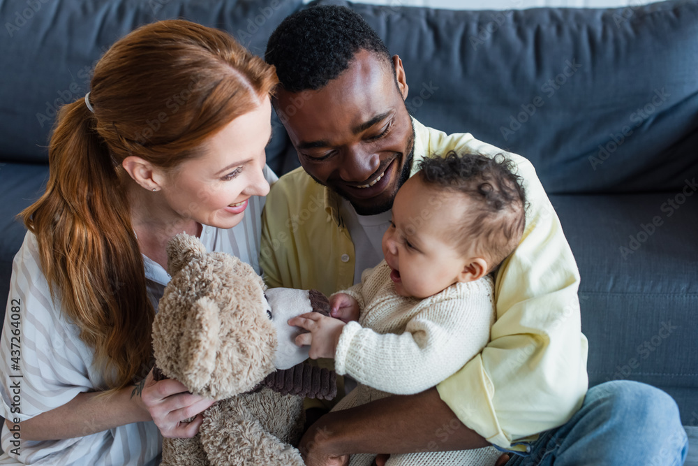 happy interracial parents playing with baby girl and teddy bear