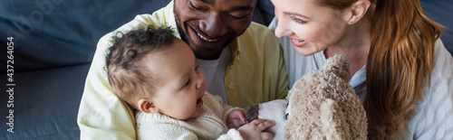 happy interracial couple looking at cheerful baby touching teddy bear, banner