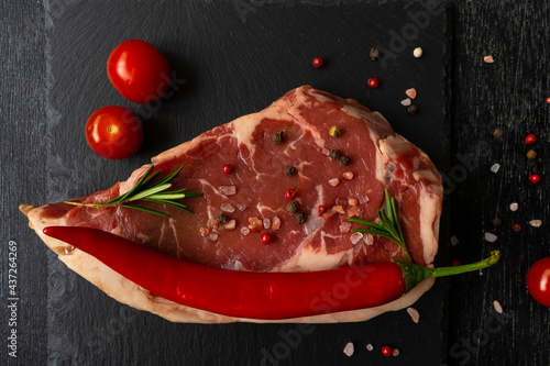 Raw piece of ribeye beef meat on a slate board r on a wooden table stacked tomatoes, hot pepper smile,spice and himalayan salt. Fresh food. Everything for a barbecue.