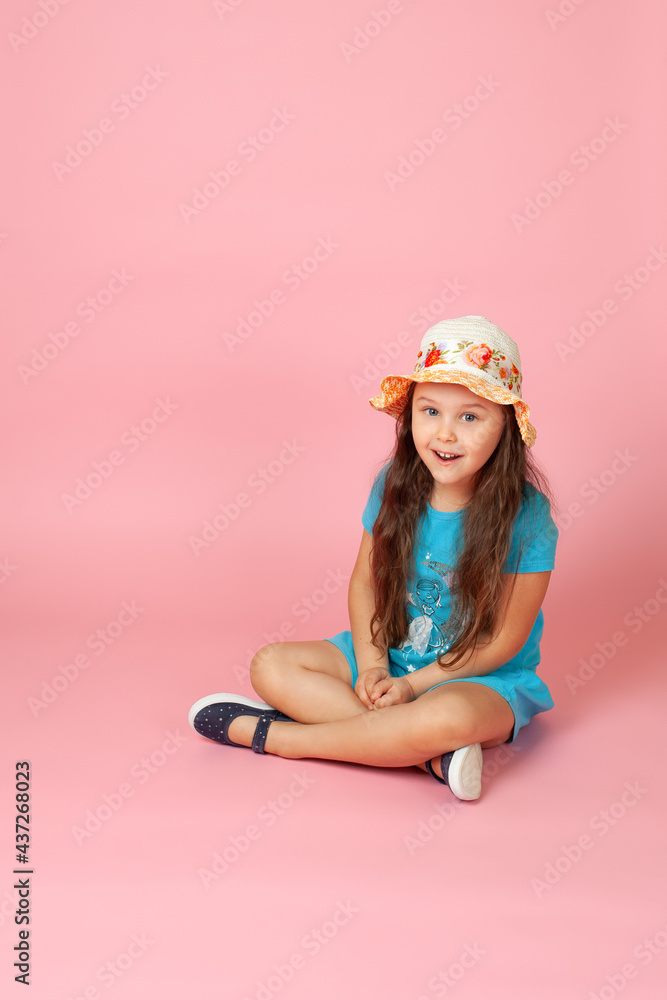 full-length portrait of a doubting, shocked girl with long wavy hair in a straw hat and blue dress sitting cross-legged on the floor, isolated on a pink background. full-length portrait of a doubting, shocked girl with long wavy hair in a straw hat and blue dress sitting cross-legged on the floor, isolated on a pink background.