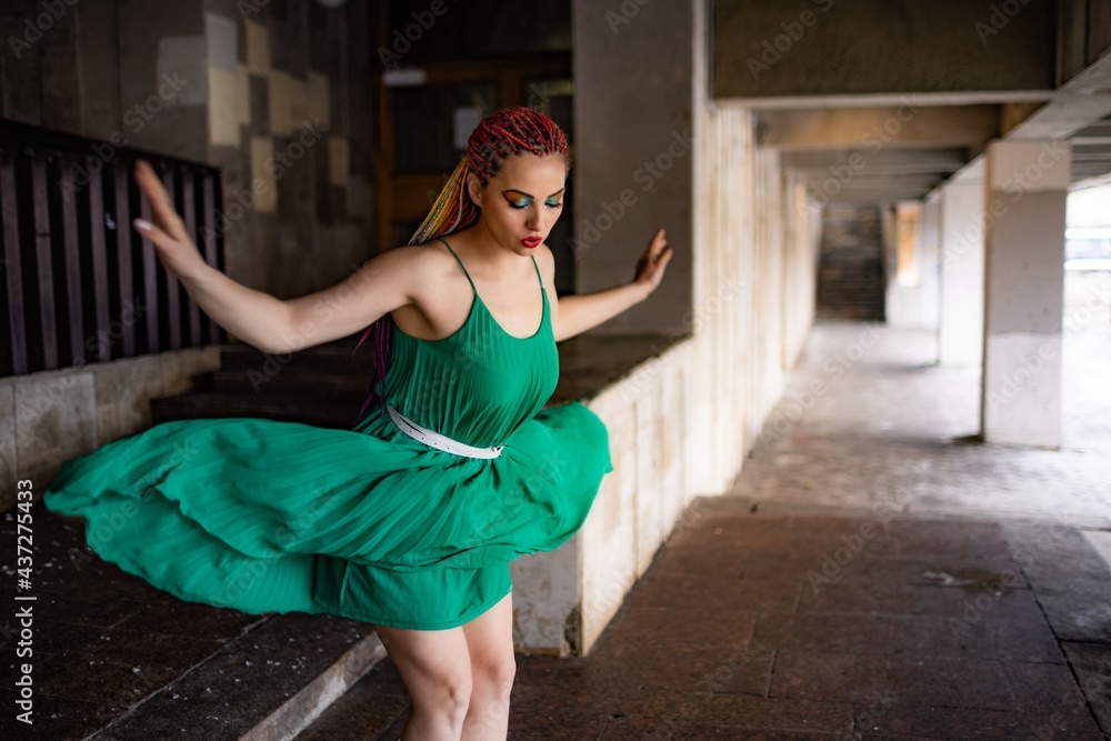 Fototapeta premium A girl with bright braids and glitter colored makeup in a bright green spring dress jumps off the steps of an old building