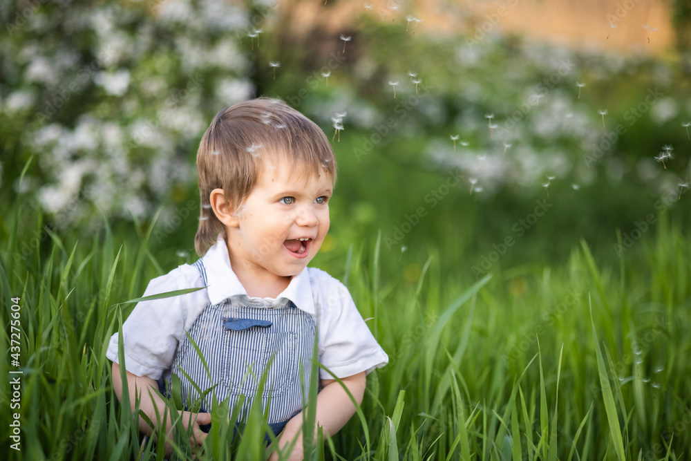Cute boy in fashionable clothes with blue eyes plays and jumps in the tall grass in a large green blooming garden Cute boy in fashionable clothes with blue eyes plays and jumps in the tall grass in a large green blooming garden