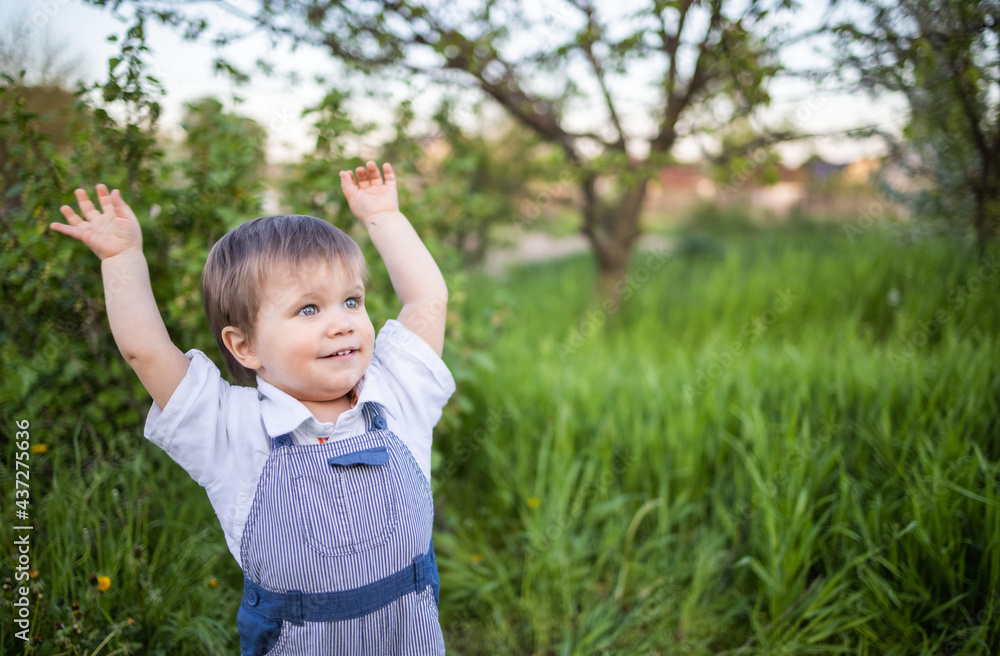 Cute boy in fashionable clothes with blue eyes plays and jumps in the tall grass in a large green blooming garden Cute boy in fashionable clothes with blue eyes plays and jumps in the tall grass in a large green blooming garden