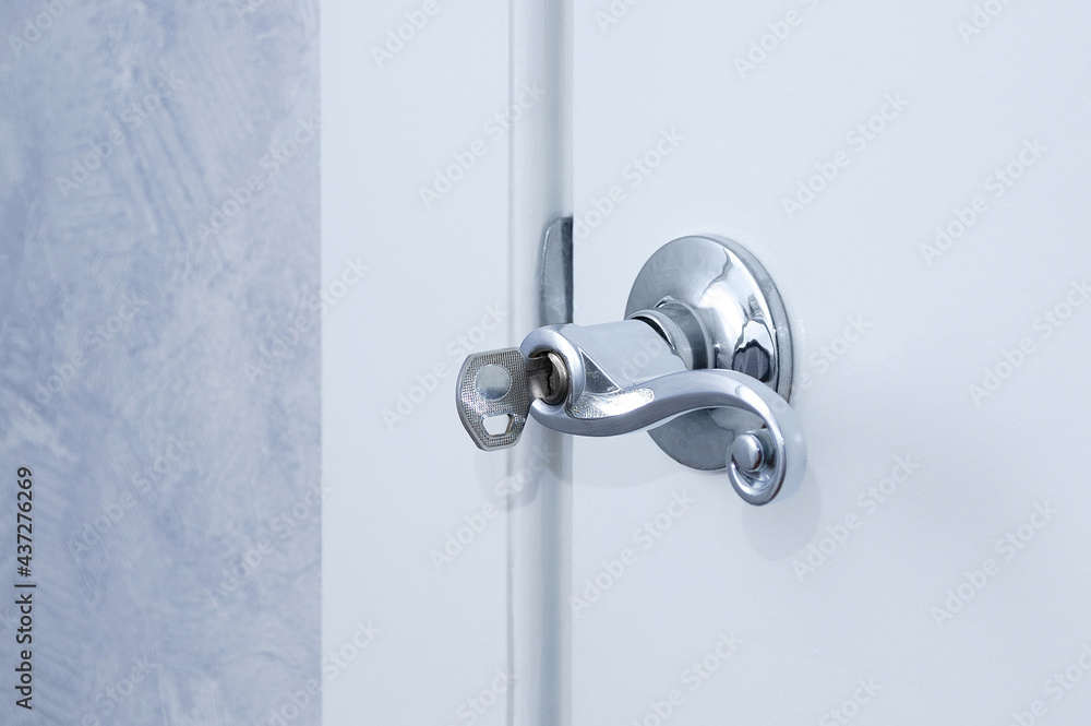 Metallic gray handle with key on a white closed door in a room. Metallic gray handle with key on a white closed door in a room.