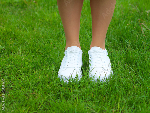 Legs of a girl in white sneakers on green grass (lawn). Close up of a pair of white shoes on the legs of a girl against a background of green grass.