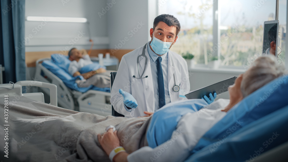 Fototapeta premium Hospital Ward: Friendly Doctor Wearing Face Mask Talks to Beautiful Senior Female Patient Resting in Bed, Explains Test Results, Gives Recovery Advice. Physician and Old Lady Recovering after Covid-19