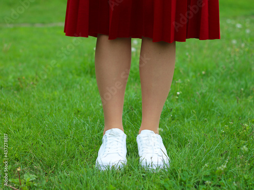 Legs of a girl in white sneakers on green grass (lawn). Close up of a pair of white shoes on the legs of a girl against a background of green grass.