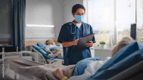 Hospital Ward  Friendly Nurse Wearing Face Mask Talks to Beautiful Senior Female Patient Resting in Bed  Checks Her Vitals  Writes down Information. Head Nurse and Old Lady Recovering after Covid-19