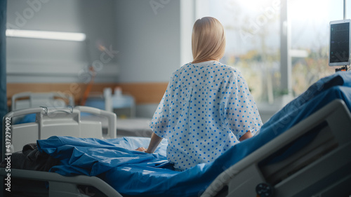 Hospital Ward: Beautiful Caucasian Female Patient Sitting on a Bed, Fully Recovering after Successful Surgery, Covid-19, Sickness she Looks Around and out of the Window with a Beautiful Sunny View photo