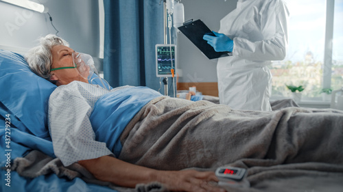 Fotografie Hospital Coronavirus Emergency Department Ward: Doctor wearing Coverall Writes Down Vitals of a Senior Female Patient with Oxygen Mask Resting in Bed