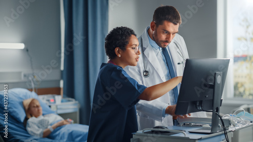 Hospital Ward: Caucasian Doctor Talks With Professional Black Head Nurse, They Use High-Tech Computer. In Background Patient in Bed Recovering after Successful Surgery. Health Care Specialists