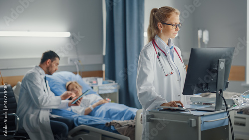 Hospital Ward  Professional Female Doctor or Surgeon Uses Medical Computer. In the Background Modern Equipment Clinic Doctor Sitting and Caring for Patient Recovering After Successful Surgery in Bed