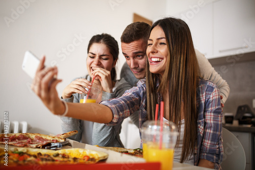 Group of young students eating pizza at the home party.Thay taking selfi with smart phone. 