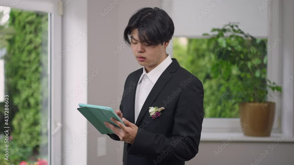 Smiling Asian groom messaging online standing indoors in elegant wedding suit. Portrait of positive happy young man texting on tablet surfing social media on marriage morning Smiling Asian groom messaging online standing indoors in elegant wedding suit. Portrait of positive happy young man texting on tablet surfing social media on marriage morning
