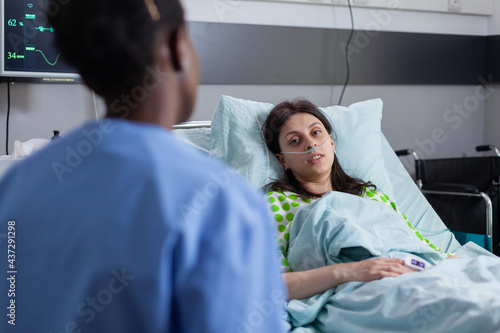 Hospitalized sick woman lying in bed while afro-american nurse checking disease symptom writing medical expertise on clipboard. Assistant discussing illness therapy working in hospital ward