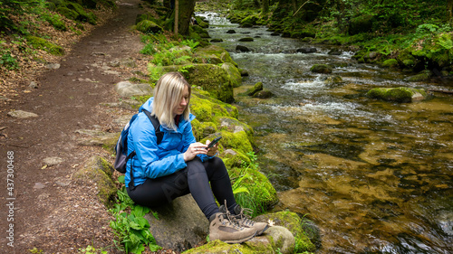 Woman in hiking boots sitting by the picturesque forest mountain stream and typing in her smart phone.